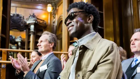 Ben Gregory-Ring/Tring Park School Emeka Sesay smiling with other people in a room, which looks like a historic hall with wood panelling. They are all looking off-camera to the left and some are clapping. He is wearing sunglasses, a grey jumper and beige coat.