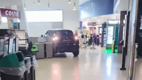 A black car reverses inside a service station, between Costa and WH Smith