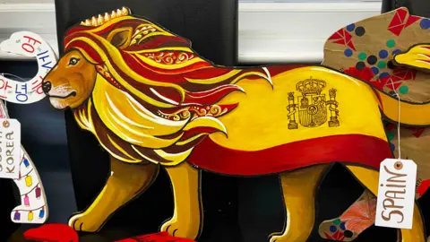 A lion is painted in traditional yellow and red colours, with a label attached saying "Spain"