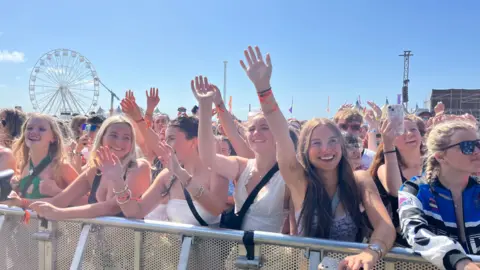 A shot of the crowd at Boardmasters festival. It shows a row of seven women against the barrier with their hands in the air in sunny weather. 