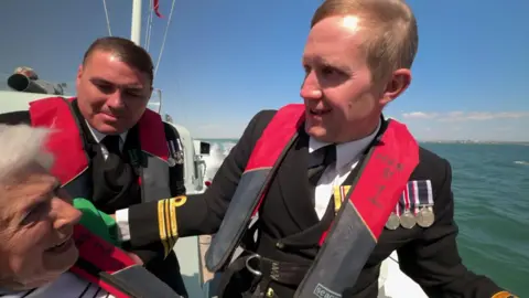 Dorothy is on board the gun boat, wind blowing her white hair, beaming. She's flanked by two navy officers. Everyone is wearing life jackets.