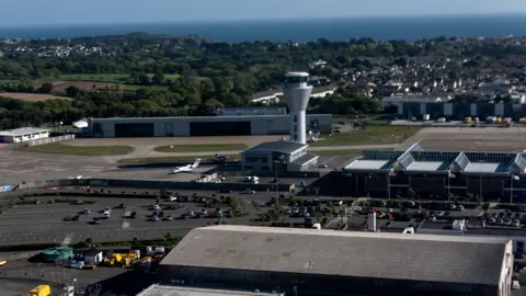 BBC Aerial view of Jersey airport. A large car park, next to the airport, is half filled with vehicles. A wall separates the car park from the airport. Large buildings and an air traffic control tower overlook the car park. A plane is parked at the airport. Green trees and residential homes can be seen in the distance behind the site.