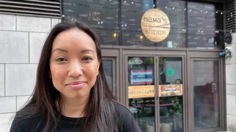 Woman with long dark hair stands in front of business front door with Mama's Kitchen sign in wood.