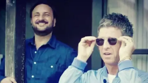 Nick Brine and Noel Gallagher. Noel is wearing sunglasses, a blue jacket and a gold chain bracelet. Nick has dark hair and a beard and is wearing a dark blue shirt. 