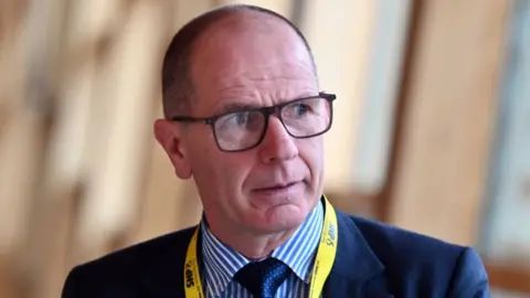 Getty Images Jim Fairlie, who is bald with glasses, in a close-up shot. He is wearing a dark blue suit and tie with a blue and white striped shirt. He is wearing a yellow SNP lanyard around his neck. 