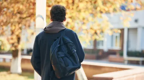 A student walks away from the camera. You can't see his face, but he has brown hair and wears a hoodie with a grey jacket and a black backpack.