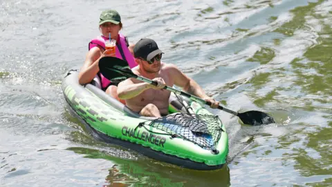 Two people kayak along the River Cam. One holds an oar while the other holds a cold drink.