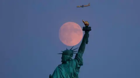 Getty Images A plane flying above the Statue of Liberty with a full moon in the background