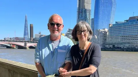 Karen Goddard Mr and Mrs Goddard stand hand in hand along the River Thames in London on a sunny day. They both wear sunglasses. Large glass buildings can be seen behind them including the Shard and a bridge.