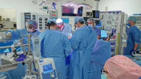 A large medical team in full surgical attire works together in an operating room. Multiple people stand around an operating table, surrounded by monitoring equipment, surgical tools, and machines displaying vital signs. Bright overhead surgical lights illuminate the centre of the room, and medical monitors show detailed images related to the procedure. The room is filled with specialised equipment and supplies arranged on carts and stands.
