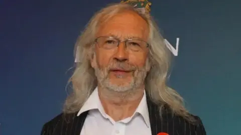 LDRS An image of Phil Riley. He has long hair and is wearing a black striped suit and white shirt.