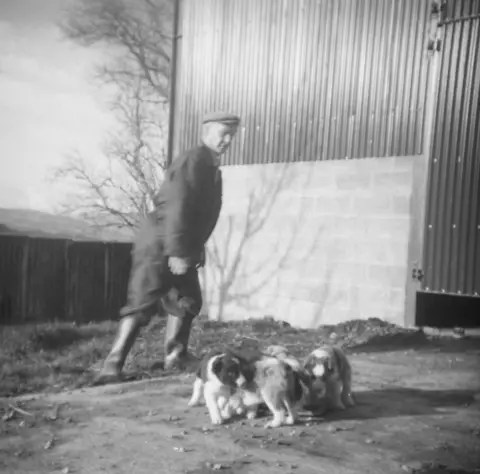 Family photo A man in a boiler suit and a flat cap stands outside a corrugated metal barn wall looking down on a litter of puppies
