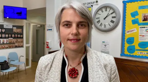 A woman with grey short hair wearing a light grey jacket and red flower pendant.
