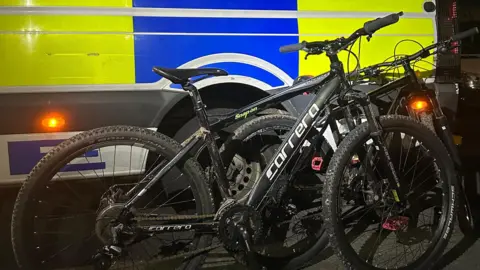 Two black bicycles have been propped up next to a yellow and blue police van. Both bikes are made by Carrera and have muddy wheels and mud on their frames.