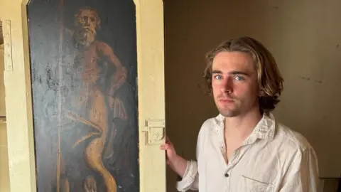 A man with brown wavy hair and a serious expression is wearing a off-white creased shirt. He is standing next to a sepia-coloured painting of an old man in a thick off-white cupboard door frame.