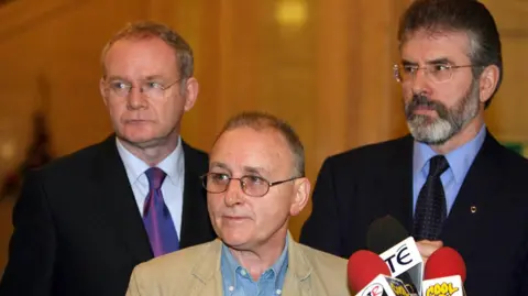 PA Media Martin McGuinness, Denis Donaldson and Gerry Adams are all looking to their right. All three are wearing glasses. Mr McGuinness and Mr Adams are both wearing dark suits, shirts and ties. Mr Donaldson is wearing a tanned coloured jacket and a denim shirt. There are three microphones in front of them.