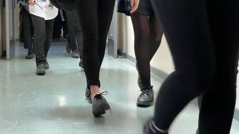 The photo shows the legs of secondary school children walking down a school corridor along a grey vinyl floor.