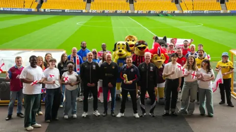 Wolves Foundation A group of people wearing football kits and holding England flags smile at the camera while standing near the pitch at Molineux Stadium.