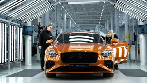Bentley An orange car on a production line. Two women are standing next to the car - one on either side. One has the door open and is looking inside.