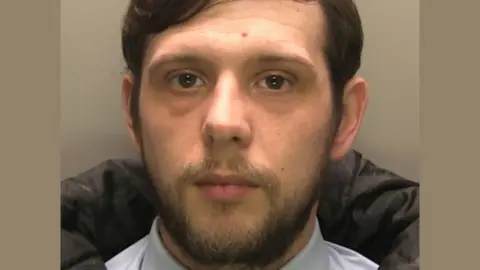 Police mugshot of a man with dark wavy hair and a short beard. He is wearing a shirt and tie and padded coat and is looking straight at the camera.