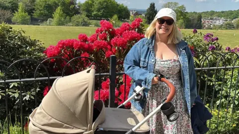 Fia Davidsson-Clayton A woman in a dress and blue jacket, sunglasses and white cap with her right hand on the handle of a cream push chair. She is stood in front of some black railings around a park
