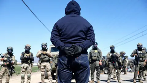 A protester stands across from immigration authorities dressed in military-SWAT uniforms. Some are holding long guns and all have on gas masks. 
