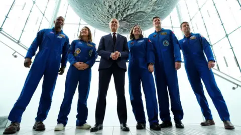Mark Carney in a suit with five astronauts in blue overalls with a moon above them. 