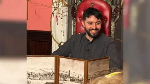 Toby Tyrrell-Jones, professional dungeon master, sat at the head of an ornate table in a fancy chair.