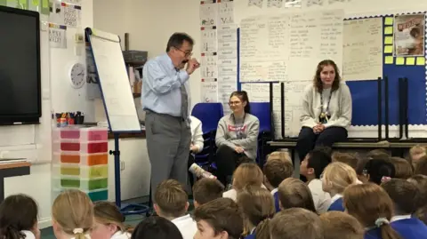 Towcester CE Primary School Lord Winston with dark hair and beard, wearing a light blue shirt and grey tie, with one fist raised. Several rows of boys and girls, some with blue school sweaters, are sitting on the floor in front of him. An adult is sitting on a desk, two others are sitting on chairs. There is a whiteboard behind Lord Winston.