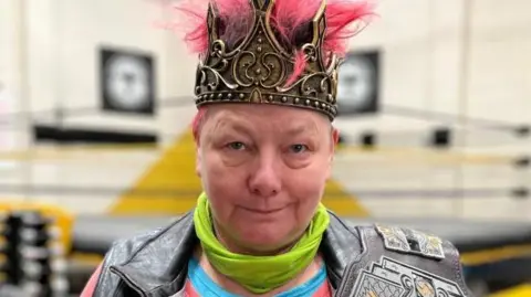Caroline Parker, an actor involved in Bardcore - Midsummer Mayhem, looking at the camera in partial costume. There is a crown on her head, and a wrestling belt over her shoulder.