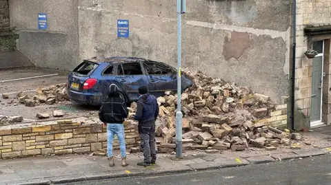 Close up of the damaged blue car, that has had its windows smashed, under stone rubble from the house. Two men are standing on the pavement staring at the damage