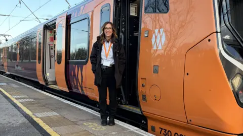 West Midlands Railway A woman with shoulder-length brown wavy hair standing on a platform next to an orange West Midlands Railway train, which has its doors open. She is smiling. She is wearing a black jacket and trousers, blue shift and orange lanyard. The sky above the train is blue.