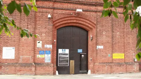 Exterior of Norwich jail. A brick building with a large wooden door and signage indicating the jail.
