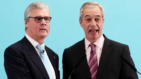 Malcolm Offord, who has grey hair and glasses, next to a smiling Nigel Farage, who has thinning grey hair. Both are wearing dark suits, and standing in front of a pale blue background. 