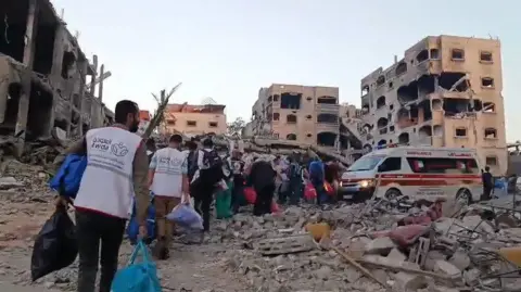 People wearing vests with the name of al-Awda hospital on the back carry bags to a row of ambulances waiting between the rubble of buildings 