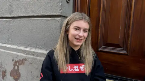 JADE kicks off UK solo tour with Belfast gig at Ulster Hall 6 A young woman sitting outside of a building, leaning between a painted grey wall and wooden doors. She has straight, brown hair with blonde highlights.