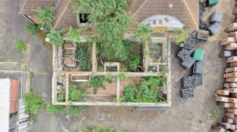 CAV Aerial An aerial of the building from the rear, showing a roofless structure overgrown with plants and weeds