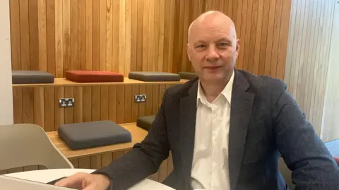 Richard Ramsey. He is bald, wearing a dark grey blazer and white shirt. He is sitting at a computer desk.