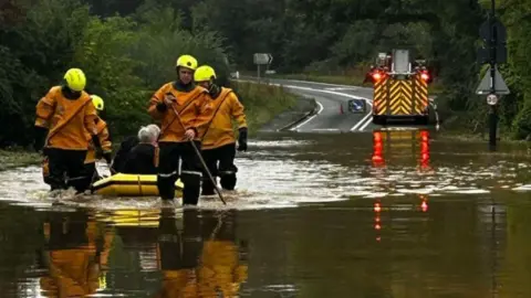 Oxfordshire Fire and Rescue Service A fire engine is seen at the edge of a heavily flooded area with fire crews in the foreground pulling a yellow boat with people onboard through the water