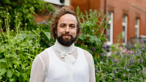 Getty Images Daf James smiles at the camera, he is wearing a white vest top and a sheer shirt over the top with a bow. He has a dark brown beard and curly brown hair. 