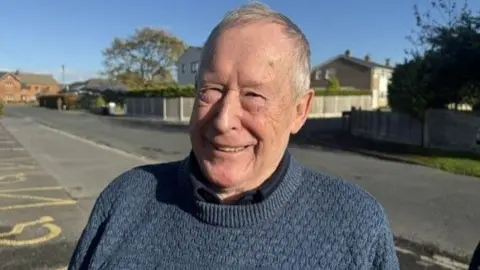 A smiling Hayden Latham: he has short white hair and wears a dark blue jumper.