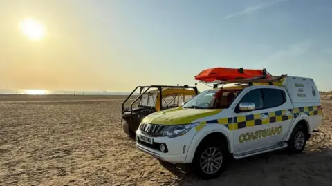 HM Coastguard Crosby Two coastguard vehicles on Crosby beach as the sun goes down over the sea. You can see some figures near the water on the sandy beach in the distance. 