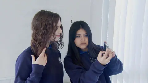 Liv and Elizabeth take a selfie on a mobile phoned, dressed in blue school jumpers. 