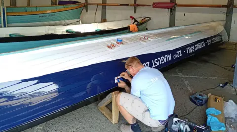 Torbay RNLI A gig boat placed upside down with a man painting it. The underside is white and the edge is being painted navy blue. The man is wearing a light blue t-shirt and stone coloured shorts with brown crocs on and black socks. He has auburn coloured short hair and has sunglasses on his head, but is faced away from the camera. There are two teal gig boats in the background.