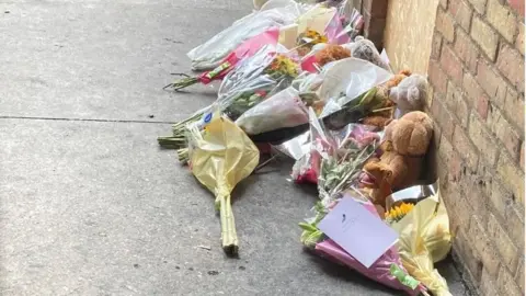 Flowers and teddy bears and lined up next to a wall on the right hand side. They are lying on a concrete floor.