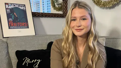 Mollie Pearce, a young woman with long, waved blonde hair, smiles at the camera sat on a sofa in a living room. She is wearing a khaki green blazer and gold necklace. Behind her is a framed copy of the Radio Times.