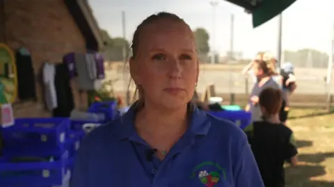 Tracey Wood in a Community Cupboard polo shirt. Uniform items are hung on a washing line and blue boxes containing clothing are seen behind her.