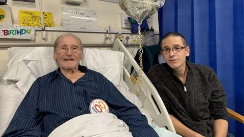 An elderly man wearing a blue shirt lying in a hospital bed. The elderly man is wearing a badge on his shirt with the number 100 on it. To the right is a man wearing a black jumper sitting in a chair. Both are looking at the camera