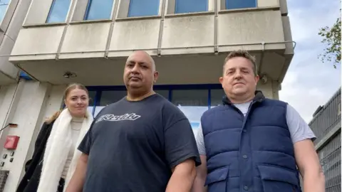 Phil Shepka/BBC Rosanna Heasman, who has blonde hair tied back, wearing a coat and white scarf; Yashpal Jassal, who is wearing a black t-shirt and Andrew, wearing a navy gilet, stood outside The Heights building