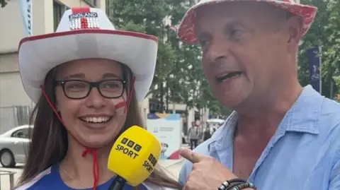 A woman and a man on a street wearing England hats and face paint with a BBC Sport microphone